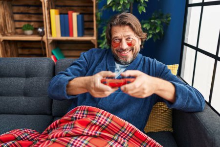 Téléchargez les photos : Homme d'âge moyen portant sous les yeux patchs jouer à un jeu vidéo à la maison - en image libre de droit