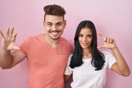 Téléchargez les photos : Jeune couple hispanique debout sur fond rose geste souriant et confiant avec la main faisant signe de petite taille avec les doigts à la recherche et la caméra. concept de mesure. - en image libre de droit