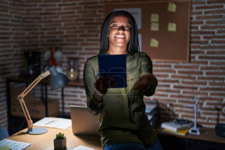 Téléchargez les photos : Jeune afro-américain avec des tresses travaillant au bureau la nuit souriant gai offrant main de paume donnant assistance et acceptation. - en image libre de droit