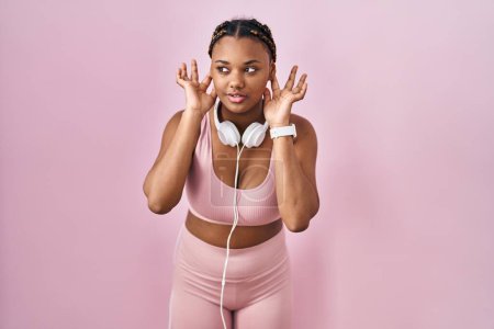 Foto de Mujer afroamericana con trenzas que usan ropa deportiva y auriculares tratando de escuchar ambos gestos en las orejas, curiosa de chismes. problema auditivo, sordo - Imagen libre de derechos
