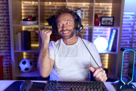 Foto de Hombre de mediana edad con barba jugando videojuegos con auriculares sonriendo con la cara feliz mirando y apuntando hacia un lado con el pulgar hacia arriba. - Imagen libre de derechos