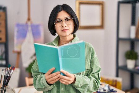 Téléchargez les photos : Young chinese woman artist reading book at art studio - en image libre de droit
