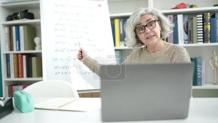Téléchargez les photos : Middle age woman with grey hair teacher teaching online maths lesson at university classroom - en image libre de droit