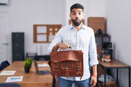 Photo for Hispanic man with beard working at the office holding briefcase scared and amazed with open mouth for surprise, disbelief face - Royalty Free Image