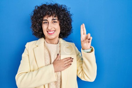Téléchargez les photos : Jeune femme brune avec les cheveux bouclés debout sur fond bleu souriant jurant avec la main sur la poitrine et les doigts vers le haut, faisant un serment de fidélité - en image libre de droit