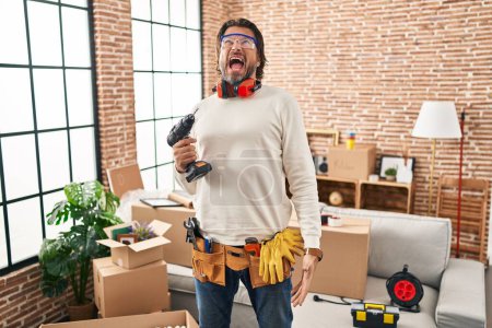 Foto de Handsome middle age man holding screwdriver at new home angry and mad screaming frustrated and furious, shouting with anger. rage and aggressive concept. - Imagen libre de derechos