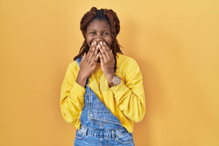Téléchargez les photos : Femme africaine debout sur fond jaune rire et rire embarrassé couvrant la bouche avec les mains, commérages et concept scandale - en image libre de droit