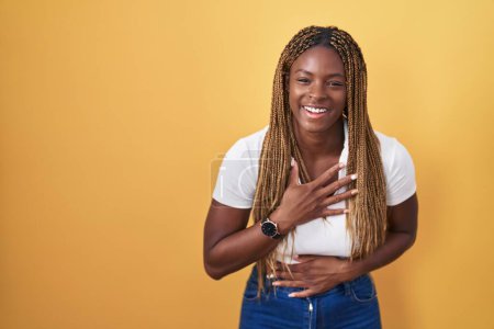 Téléchargez les photos : Femme afro-américaine avec des cheveux tressés debout sur fond jaune souriant et riant fort à haute voix parce que drôle blague folle avec les mains sur le corps. - en image libre de droit