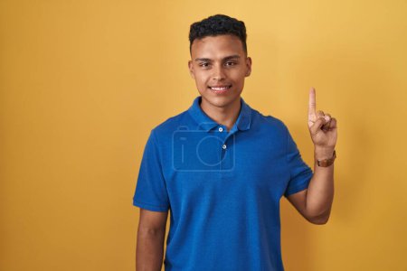 Young hispanic man standing over yellow background showing and pointing up with finger number one while smiling confident and happy. 