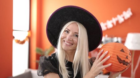Foto de Young blonde woman wearing witch costume holding halloween pumpkin basket at home - Imagen libre de derechos