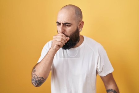 Foto de Hombre hispano joven con barba y tatuajes de pie sobre fondo amarillo sintiéndose mal y tosiendo como síntoma de resfriado o bronquitis. concepto de atención sanitaria. - Imagen libre de derechos
