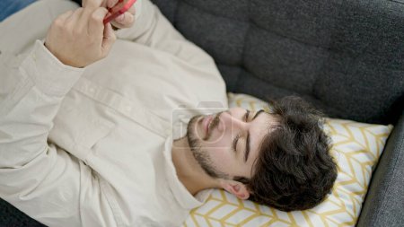 Téléchargez les photos : Jeune homme hispanique utilisant un smartphone couché sur le canapé à la maison - en image libre de droit