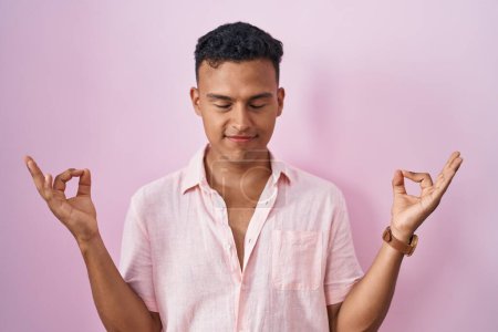 Foto de Joven hombre hispano de pie sobre fondo rosa relajado y sonriente con los ojos cerrados haciendo gesto de meditación con los dedos. concepto de yoga. - Imagen libre de derechos