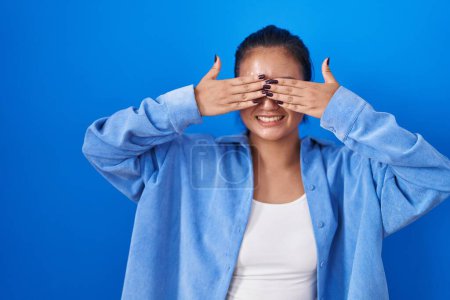 Photo for Asian young woman standing over blue background covering eyes with hands smiling cheerful and funny. blind concept. - Royalty Free Image
