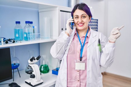 Téléchargez les photos : Plus taille femme esprit cheveux violets travaillant au laboratoire scientifique parlant sur g le téléphone souriant heureux pointant avec la main et le doigt sur le côté - en image libre de droit