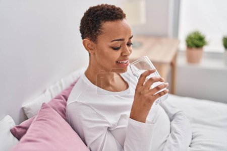 Photo for African american woman drinking glass of water sitting on bed at bedroom - Royalty Free Image