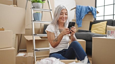 Téléchargez les photos : Femme aux cheveux gris du moyen âge utilisant un smartphone assis sur le sol à la nouvelle maison - en image libre de droit