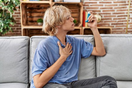 Téléchargez les photos : Jeune homme blond utilisant inhalateur assis sur le canapé à la maison - en image libre de droit