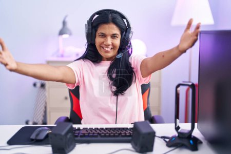 Photo for Mature hispanic woman playing video games at home looking at the camera smiling with open arms for hug. cheerful expression embracing happiness. - Royalty Free Image