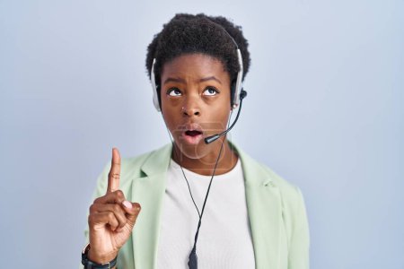 Téléchargez les photos : Femme afro-américaine portant casque agent centre d'appel étonné et surpris de regarder vers le haut et pointant avec les doigts et les bras levés. - en image libre de droit
