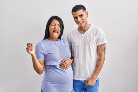 Foto de Joven pareja hispana esperando un bebé de pie sobre el fondo sacando la lengua feliz con expresión divertida. concepto de emoción. - Imagen libre de derechos