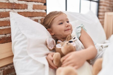 Photo for Adorable blonde girl holding doll lying on bed at bedroom - Royalty Free Image