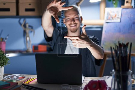 Téléchargez les photos : Jeune homme caucasien utilisant ordinateur portable la nuit au studio d'art souriant faire cadre avec les mains et les doigts avec le visage heureux. concept de créativité et photographie. - en image libre de droit
