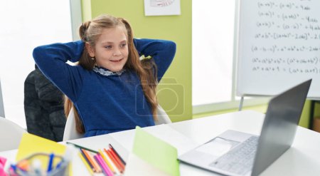 Foto de Adorable estudiante rubia chica usando portátil descansando con las manos en la cabeza en el aula - Imagen libre de derechos