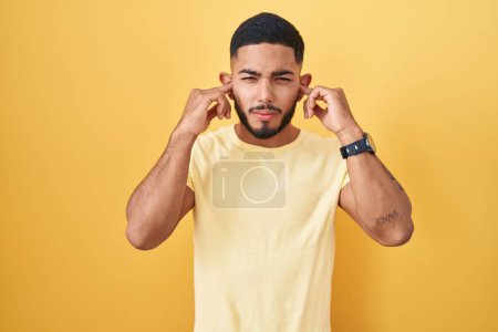 Photo for Young hispanic man standing over yellow background covering ears with fingers with annoyed expression for the noise of loud music. deaf concept. - Royalty Free Image
