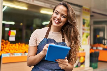 Photo for Young beautiful hispanic woman waitress smiling confident using touchpad at fruit store - Royalty Free Image
