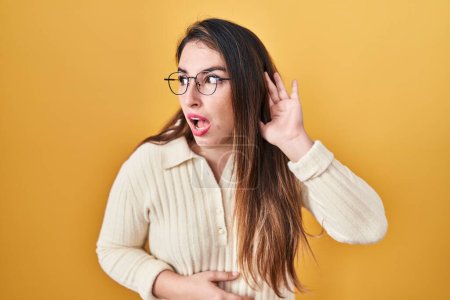 Photo for Young hispanic woman standing over yellow background smiling with hand over ear listening an hearing to rumor or gossip. deafness concept. - Royalty Free Image
