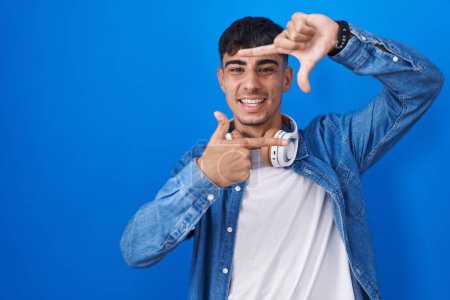 Téléchargez les photos : Jeune homme hispanique debout sur fond bleu souriant faisant cadre avec les mains et les doigts avec le visage heureux. concept de créativité et photographie. - en image libre de droit