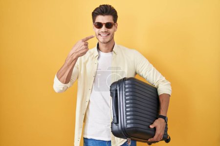 Téléchargez les photos : Jeune homme hispanique tenant une valise allant en vacances d'été pointant du doigt vers le visage et le nez, souriant gai. concept de beauté - en image libre de droit
