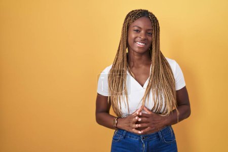 Téléchargez les photos : Femme afro-américaine avec les cheveux tressés debout sur fond jaune mains ensemble et doigts croisés souriant détendu et joyeux. succès et optimisme - en image libre de droit