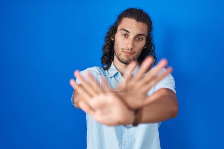 Foto de Joven hombre hispano de pie sobre fondo azul expresión de rechazo cruzando brazos y palmas haciendo signo negativo, cara enojada - Imagen libre de derechos
