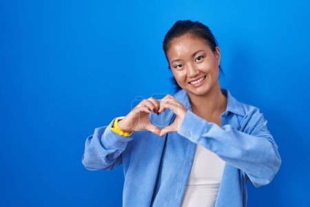 Téléchargez les photos : Asiatique jeune femme debout sur fond bleu souriant dans l'amour faisant forme de symbole de coeur avec les mains. concept romantique. - en image libre de droit