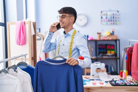 Téléchargez les photos : Jeune homme hispanique tailleur parlant sur smartphone tenant t-shirt à la boutique de tailleur - en image libre de droit