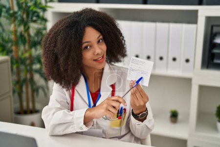 Téléchargez les photos : Young african american woman wearing doctor uniform holding covid-19 vaccination record card at clinic - en image libre de droit
