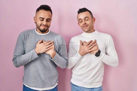 Téléchargez les photos : Couple homosexuel debout sur fond rose souriant avec les mains sur la poitrine avec les yeux fermés et geste reconnaissant sur le visage. concept de santé. - en image libre de droit