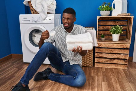 Foto de Hombre afroamericano sosteniendo ropa limpia y polvo de lavandería expresión despistada y confusa. concepto de duda. - Imagen libre de derechos