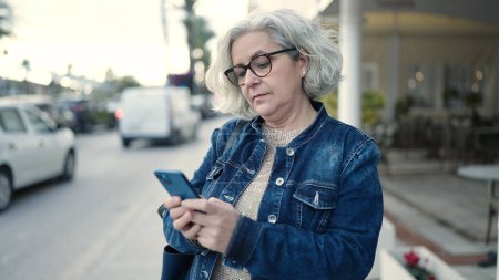 Foto de Middle age woman with grey hair using smartphone at street - Imagen libre de derechos