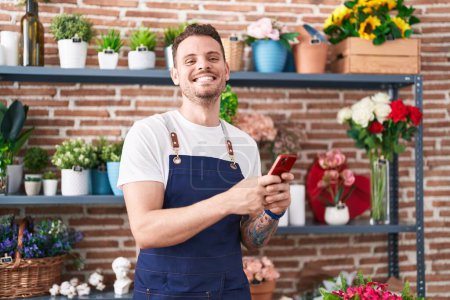 Téléchargez les photos : Jeune homme hispanique fleuriste souriant confiant en utilisant un smartphone chez le fleuriste - en image libre de droit