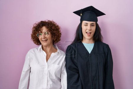 Foto de Madre e hija hispanas con gorra de graduación y bata de ceremonia guiñando el ojo mirando a la cámara con expresión sexy, cara alegre y feliz. - Imagen libre de derechos