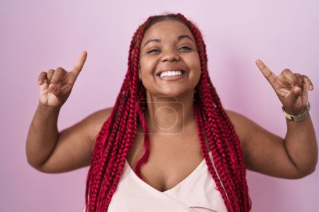 Téléchargez les photos : Femme afro-américaine aux cheveux tressés debout sur fond rose souriant étonné et surpris et pointant vers le haut avec les doigts et les bras levés. - en image libre de droit
