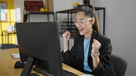 Téléchargez les photos : Jeune belle ouvrière hispanique utilisant un ordinateur portable travaillant avec l'expression gagnante au bureau - en image libre de droit