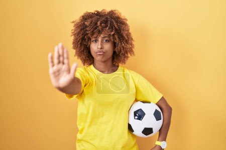 Téléchargez les photos : Jeune femme hispanique avec les cheveux bouclés tenant ballon de football avec la main ouverte faisant stop signe avec expression sérieuse et confiante, geste de défense - en image libre de droit