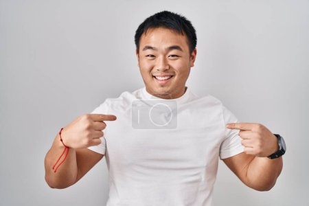 Foto de Joven hombre chino de pie sobre fondo blanco mirando confiado con sonrisa en la cara, señalándose con los dedos orgullosos y felices. - Imagen libre de derechos