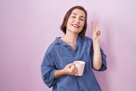 Téléchargez les photos : Femme hispanique du Moyen Age buvant une tasse de café souriant en regardant la caméra montrant les doigts faisant signe de victoire. numéro deux. - en image libre de droit