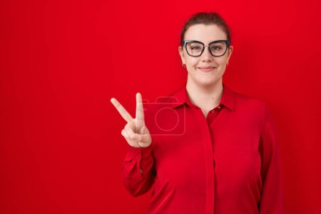 Téléchargez les photos : Jeune femme hispanique avec les cheveux rouges debout sur fond rouge montrant et pointant vers le haut avec les doigts numéro deux tout en souriant confiant et heureux. - en image libre de droit