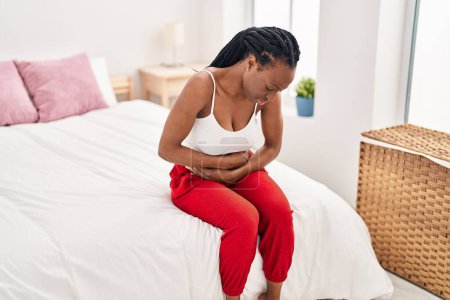 Foto de African american woman suffering for menstrual pain sitting on bed at bedroom - Imagen libre de derechos